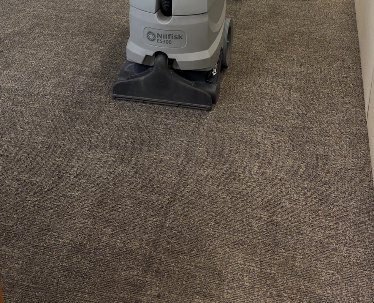 Lavagem Carpete em Placas por Extração Pessoa utilizando uma extratora profissional Nilfisk ES300 para a lavagem profunda de um carpete modular marrom em um ambiente comercial. A máquina está centralizada, mostrando o processo de lavagem do carpete por extração.
